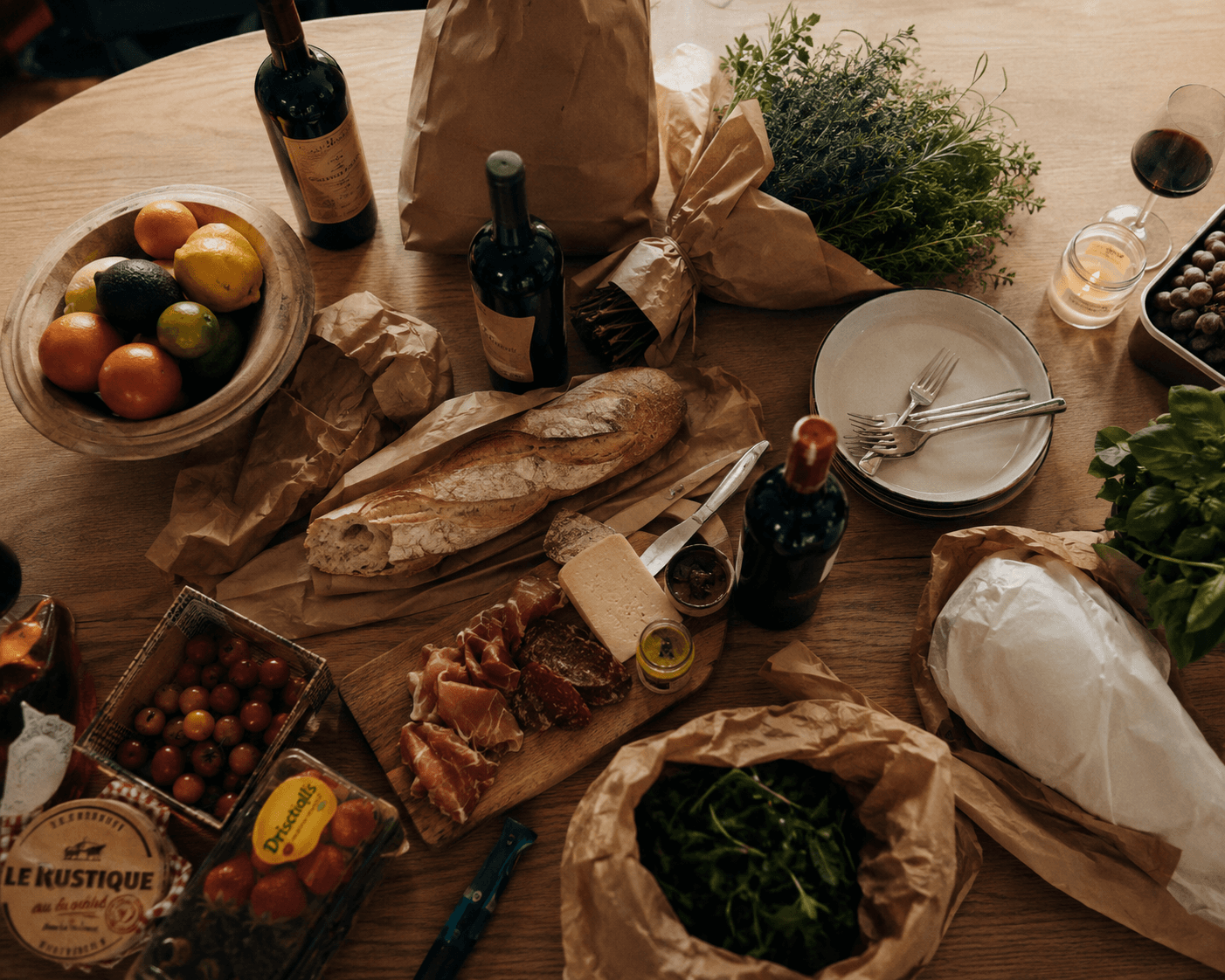 Table ronde avec bouteilles de vin, baguette, charcuterie et courses encore dans leurs sacs kraft — chacun a apporté sa part
